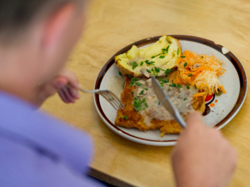 Mensch sitzt vor einem Teller mit Ofenkartoffel, Karottensalt und Soja-Schnitzel