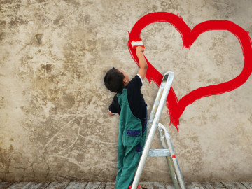 Kind malt ein Herz mit roter Farbe an eine Hauswand.