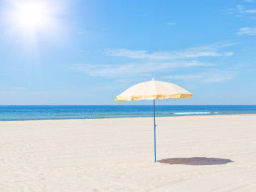 Blick auf einen glühendheißen Sandstrand mit gelben Sonnenschirm und blauem Meer.