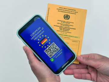 Handy mit europaweit anerkanntem Nachweis des Impfstatus und gelbes Impfheft im Hintergrund