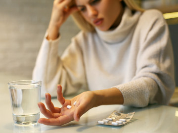 Frau mit Kopfschmerzen sitzt vor einem Wasserglas und hat zwei Tabletten in der Hand

