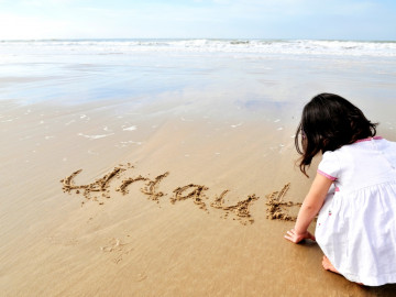 kleines Mädchen hockt am Strand und schreibt das Wort Urlaub in den Sand