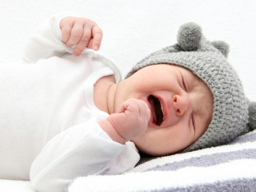Weinendes Baby mit weißem Body und Bommelmütze.