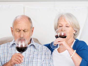 Ein älterer Mann und eine ältere Frau sitzen auf der Couch und trinken Rotwein.