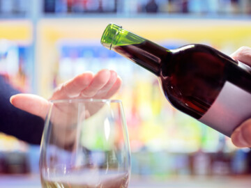 Weinflasche aus der in ein Glas Wein gegossen werden soll. Eine Hand legt sich abwehrend über das Glas.