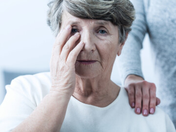 Seniorin hält sich die Hand vors rechte Auge. Frau hinter ihr legt ihre Hand auf die Schulter der Seniorin