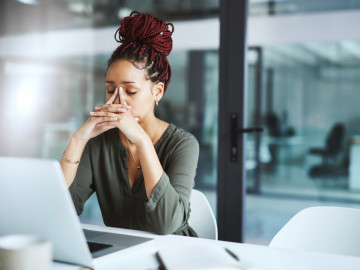 Gestresste Frau sitzt mit Fingern an der Nase am Laptop