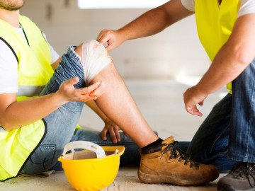 Verletzter Bauarbeiter mit verbundenem Knie wird von einem Kollegen betreut 