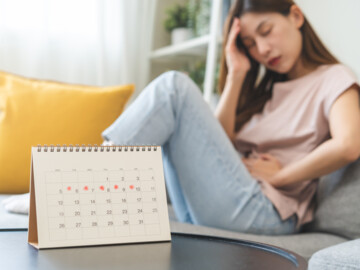 Junge Frau mit Bauch- und Kopfschmerzen auf dem Sofa im Hintergrund. Im Vordergrund ein Kalender mit fünf rot markierten Tagen.