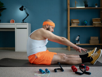 Mann mit weißem Unterhemd und orangefarbener Sporthose und Stirnband versucht sitzend seine Fußspitzen zu erreichen.