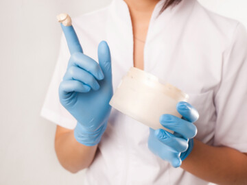 Frau mit blau-behandschuhter Hand hat Salbe auf dem Zeigefinger und eine Cremedose in der Hand