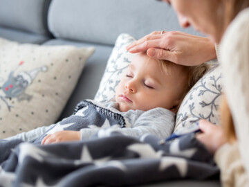 Mutter fühlt die Stirn bei ihrem kleinen Kinde, das mit geschlossenen Augen und zugedeckt auf dem Sofa liegt.