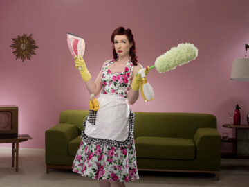 Hausfrau im Retrolook mit Schürze und Kleid, Staubwedel und Handfeger und Schaufel in der Hand.