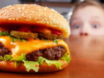 Hungriger Junge mit weit aufgerissenen Augen starrt auf einen Cheeseburger (scharf im Vordergrund)
