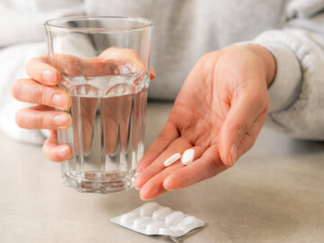 Frau hält Wasserglas in der einen Hand, in der anderen zwei Tabletten. Auf dem Tisch ein Blister mit Tabletten.