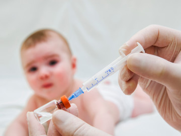 Kleinkind im Hintergrund, im Vordergrund wird eine Spritze mit Impfstoff aufgesogen