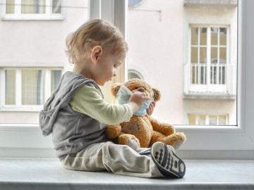 Kleines Kind sitzt mit Teddy im Arm auf der Fensterbank