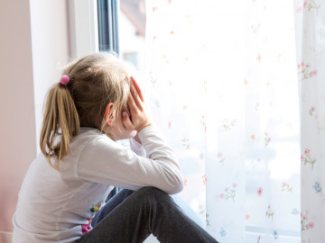 Mädchen sitzt mit Händen vor dem Gesicht am Fenster