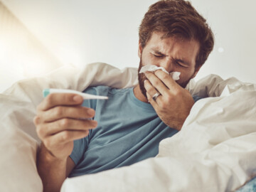 Mann mit Fieberthermometer in der Hand liegt im Bett und putzt sich die Nase
