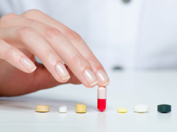 Hand hält rot-weiße Kapsel. Diese steht mittig in einer Reihe von Tabletten.