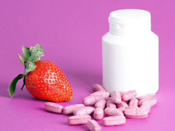Weiße Kapseldose ohne Aufschrift, eine Erdbeere sowie rosafarbene Tabletten mit Bruchkerbe im Vordergrund
