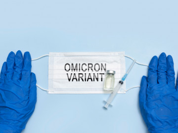 OP-Maske mit Aufdruck Omicron Variant sowie Vial und Spritze wird von zwei blau behandschuhten Händen gehalten