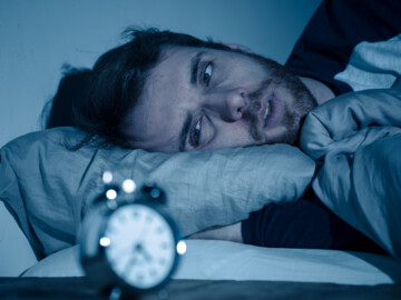 Symbolbild Schlafstörungen: Mann im Bett schaut auf Wecker.