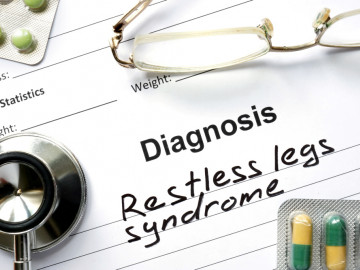 Zettel mit Diagnos „Restless less syndrome“, daraufliegend eine Lesebrille, ein Stethoskop und zwei Blister mit Tabletten und Kapseln