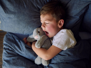 Kleiner Junge mit Kuschelhasen im Arm liegt mit geschlossenen Augen im Bett und gähnt