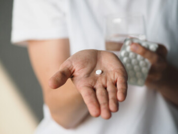 Frau hält Hand in die Kamera mit einer Tablette auf der Handinnenfläche. In der anderen Hand hält sie ein Glas mit Wasser und einen Tablettenblister