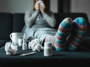 Frau auf dem Sofa, im Vordergrund genutzte Taschentücher, Nasenspray, Tablettendose und Teebecher