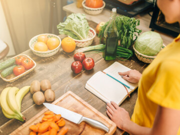 Obst und Gemüse auf einem Tisch, Frau schaut in eine Notizbuch und will kochen