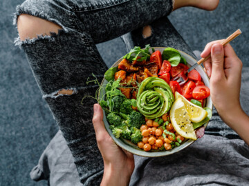 Frau in Jeans hat eine Bowl auf dem Schoß mit Brokkoli, Kichererbsen, Tomaten, Avocado und zwei Zitronenscheiben.