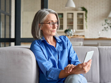 Frau um die 60 Jahre sitzt auf dem Sofa und schaut ein Video auf ihrem Handy