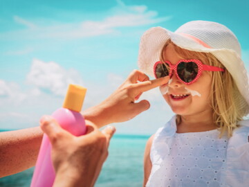 Kind mit Sonnenbrille und Sonnenhut bekommt Sonnencreme im Gesicht aufgetragen.